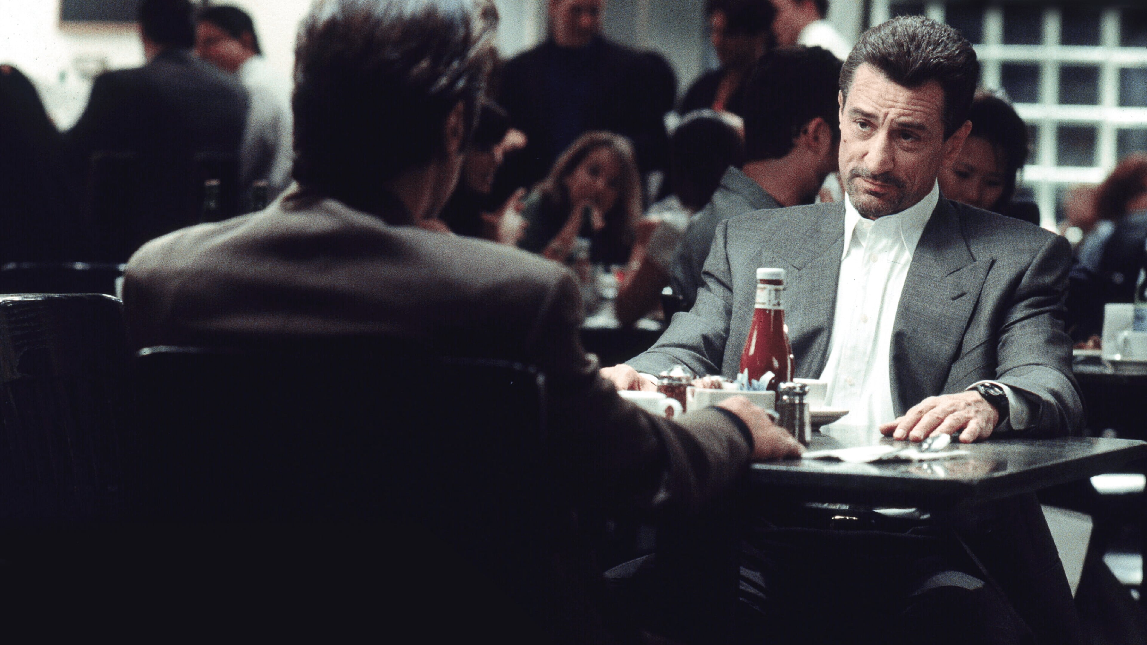 Screencap from the movie Heat showing two middle aged men in suits sitting opposite of each other in a restaurant. One of them has his back turned to the camera and we can't see his face.