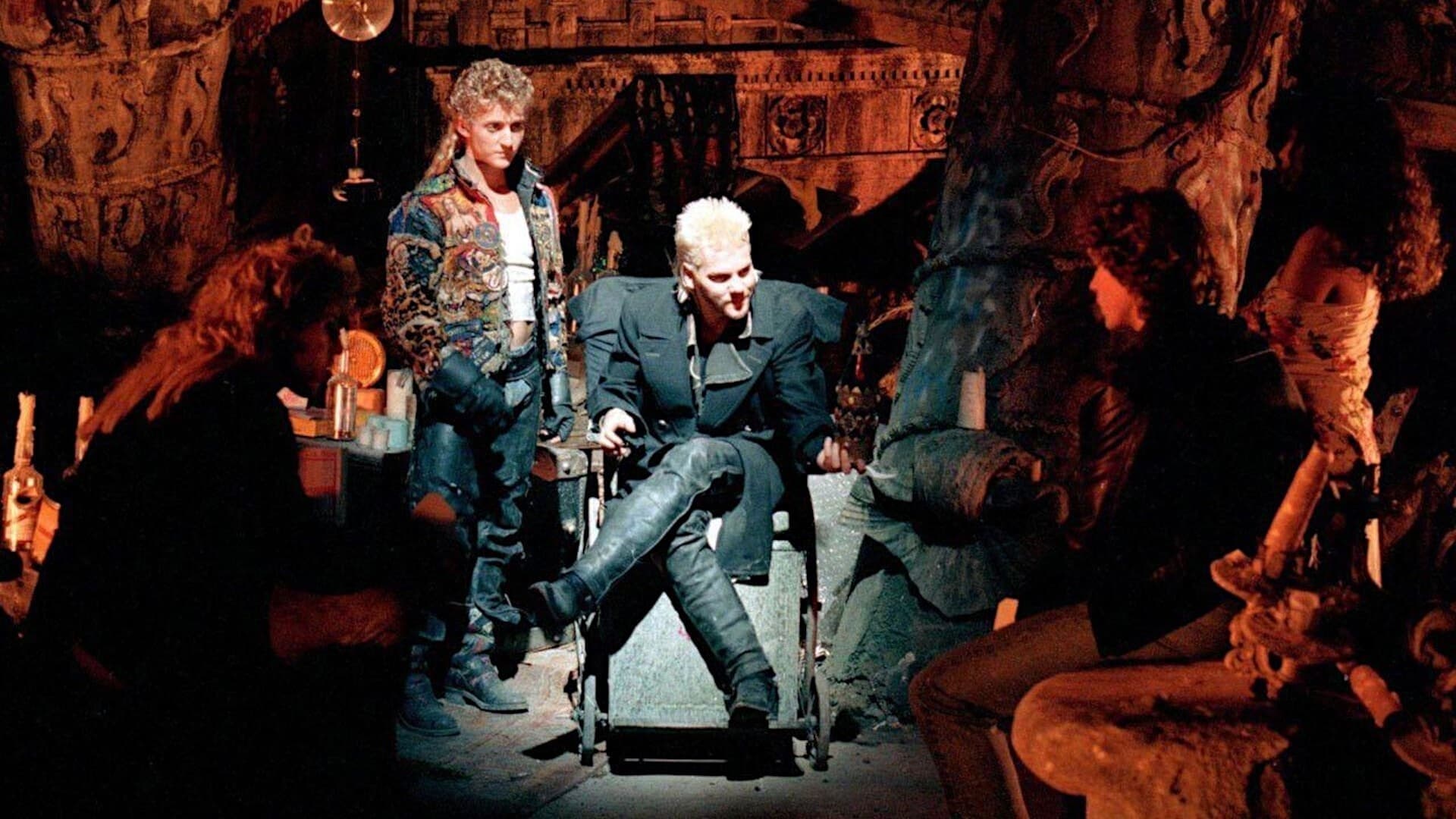 Screencap from the movie The Lost Boys of several young men with mullets and 80s hairstyles in extravagant cothing sitting in a cave. They are gathered around one of them who is illuminated by light.