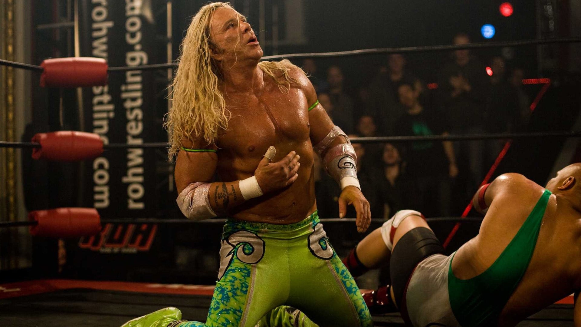 Screencap from the Movie The Wrestler showing a middle aged man with long blode hair, tanned skin and green wrestling gear in the middle of a wrestling ring. He is holding his sternum and looking up. His opponent is laying next to him on the mat in the backgrund of the shot.